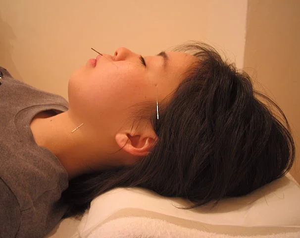 A wide-angle photo of a treatment room at an acupuncture clinic with soft evening light coming in, showing a neatly arranged treatment bed and wooden shelving